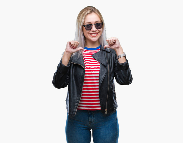 Jeune femme blonde portant une veste de mode et des lunettes de soleil sur fond isolé regardant confiant avec le sourire sur le visage, se montrant du doigt fier et heureux
. - Photo, image