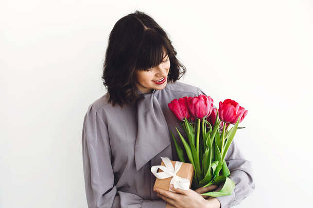 Beautiful brunette girl holding stylish bouquet of tulips and gift box on white background indoors, space for text. Happy young woman with flowers. Happy mothers day. International womens day - Photo, Image