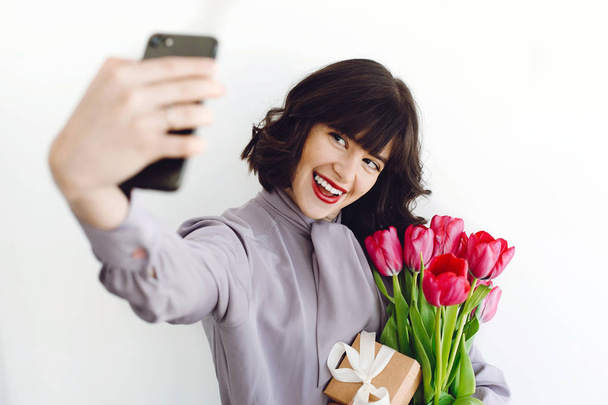 Piękna dziewczyna szczęśliwy biorąc selfie z bukietem tulipanów i pudełko na białym tle pomieszczeniu, miejsca na tekst. Stylowy, młoda kobieta z kwiatami i telefon. Międzynarodowe damskie day - Zdjęcie, obraz