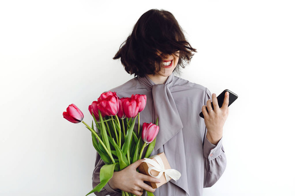 Belle fille heureuse qui s'amuse et sourit, tenant des tulipes rouges et un téléphone sur fond blanc à l'intérieur, espace pour le texte. Élégante jeune femme brune riant et agitant les cheveux. Joyeuse journée des femmes
 - Photo, image