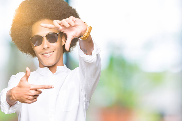 Jovem afro-americano com cabelo afro usando óculos de sol sorrindo fazendo moldura com as mãos e os dedos com rosto feliz. Conceito de criatividade e fotografia
. - Foto, Imagem