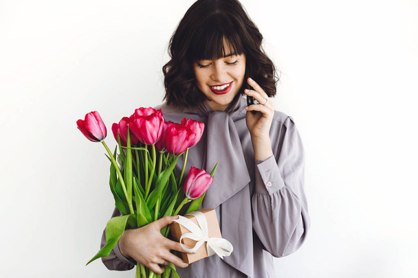Belle fille heureuse tenant des tulipes rouges, boîte cadeau et parler au téléphone sur fond blanc à l'intérieur, espace pour le texte. Jeune femme élégante tenant téléphone et fleurs. Journée internationale de la femme
 - Photo, image