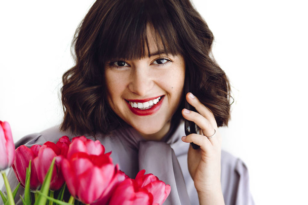 Beautiful happy girl  talking on phone and smiling, holding red tulips on white background indoors, space for text. Stylish young brunette woman with red lips and flowers. Happy womens day - Фото, зображення