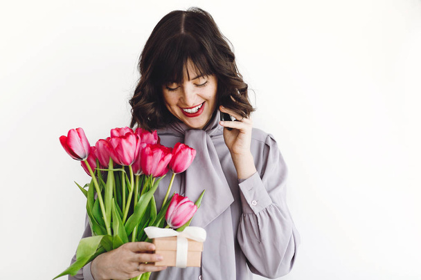 Beautiful happy girl holding red tulips, gift box and talking on phone on white background indoors, space for text. Stylish young woman holding phone and flowers.  International womens day - Photo, Image