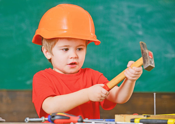 Excited kid playing with heavy hammer. Cute boy in orange helmet helping in workshop. Future profession concept - Foto, afbeelding