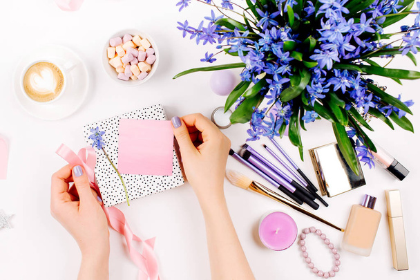 A woman unpacks a gift on background with Blue spring bouquet, coffee cup, candies and cosmetic accessories. Styled flat lay - Foto, Bild
