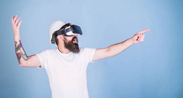 Hipster in helmet works as engineer in virtual reality. 3D design concept. Architect or engineer with virtual reality glasses. Man with beard in VR glasses pointing with finger, light blue background - Photo, image