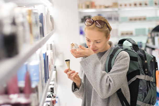 Blonďatá mladá žena cestujícím nosí cestovní batoh volba parfému v duty free obchodě letiště. - Fotografie, Obrázek