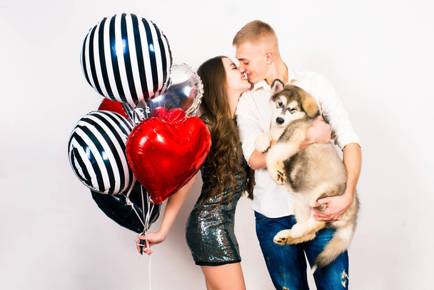Kissing couple posing on grey background with balloons hearts   and a malamute puppy.Valentine's day. - Image - Foto, Imagen