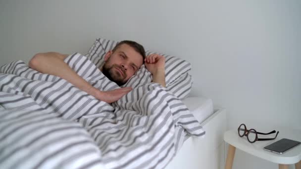 Bearded man waking up in bed under blanket, smiling, putting his eyeglasses on and taking his smartphonephone - Footage, Video