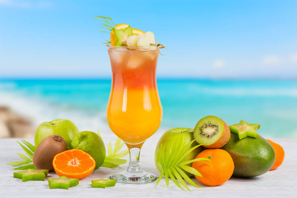 Cóctel de verano con varias frutas tropicales alrededor. Manzana, kiwi, mandarina, naranja, mango, carambola. Vaso de bebida naranja con playa borrosa en el fondo
 - Foto, Imagen