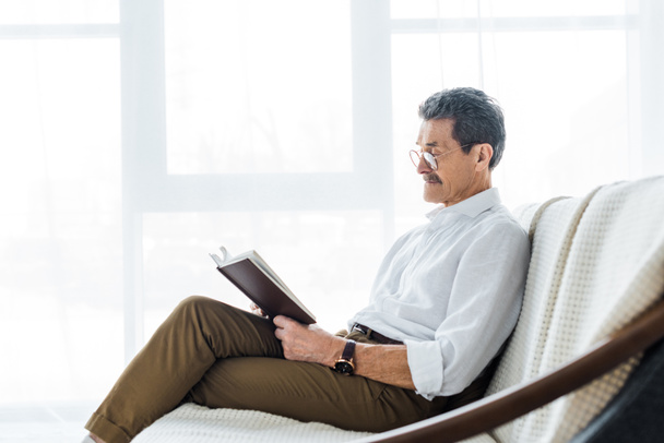 huomaavainen vanhempi mies lukee kirjaa istuessaan sohvalla
  - Valokuva, kuva