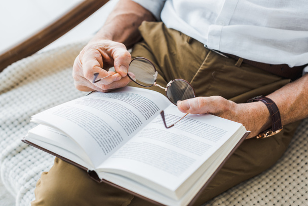 cropped view of senior man reading book and holding glasses at home - Foto, Bild