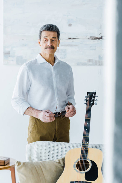 сумний старший чоловік стоїть біля акустичної гітари вдома
 - Фото, зображення