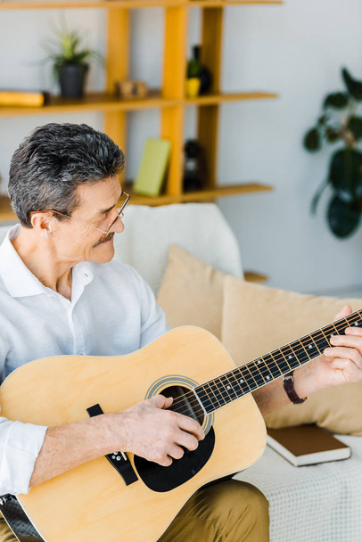 cheerful senior man playing acoustic guitar in living room - Foto, afbeelding