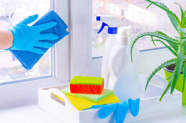 Kauçuk eldivenli elini kirli bir pencere yıkar. Kavramsal temizlik ürünleri kullanma pencere temizleme. Beyaz bir tepsi, deterjanlar ve ovma yastıkları pencere kenarında. Aloe büyüyen yakınındaki. Bulanık şehir dışında - Fotoğraf, Görsel