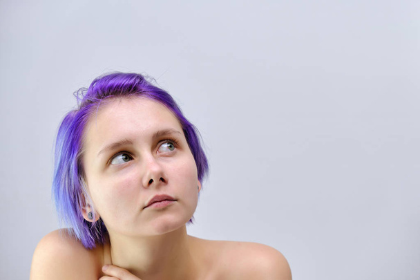 A beautiful, sexy girl with purple hair and a short haircut sits in the middle of the photo with a gray background and grimaces. She's naked and visible shoulders. - Foto, Imagem