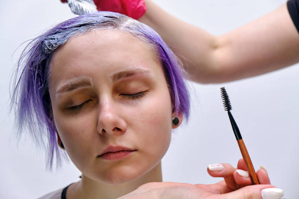 Beautiful girl with violet hair on white background master in make-up. Visible hand of the master and his makeup. The girl is in the middle of the frame and sits on a chair. - Zdjęcie, obraz