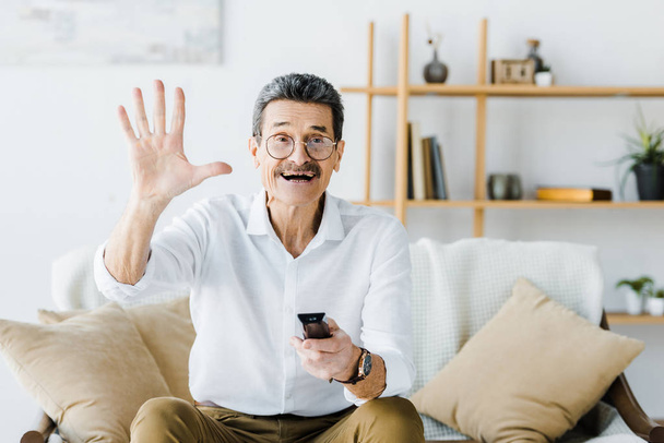 cheerful senior man sitting on sofa and holding remote control while waving hand - Fotó, kép