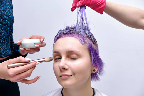 Beautiful girl with violet hair on white background master in make-up. Visible hand of the master and his makeup. The girl is in the middle of the frame and sits on a chair. - Photo, Image