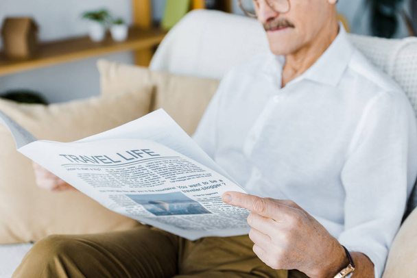 Обрезанный вид пожилого человека, читающего туристическую газету, сидя на диване
 - Фото, изображение