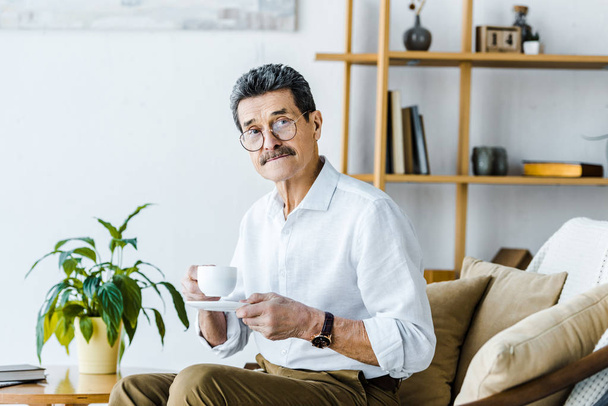 senior man holding cup with coffee while sitting on sofa  - Фото, изображение