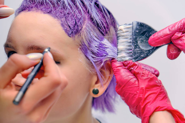 Beautiful girl with violet hair on white background master in make-up. Visible hand of the master and his makeup. The girl is in the middle of the frame and sits on a chair. - Foto, Imagen