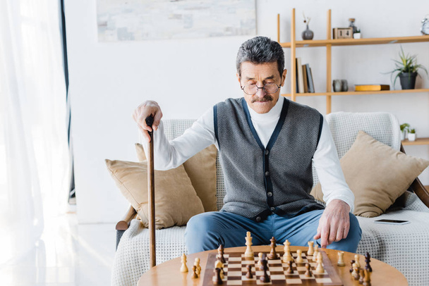 Rentner mit Schnurrbart schaut auf Schachbrett, während er zu Hause einen Gehstock hält - Foto, Bild