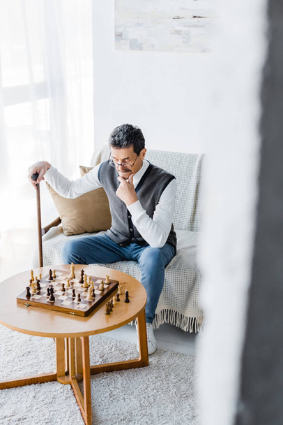 pensive retired man with mustache looking at chess board while holding walking cane at home - Photo, image