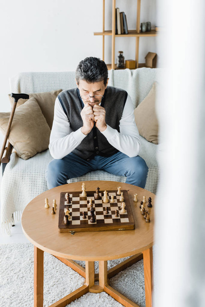thoughtful pensioner in glasses looking at chess board at home - Foto, immagini