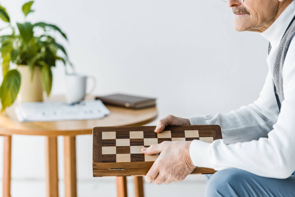 cropped view of senior man sitting and holding chess board at home - Photo, Image