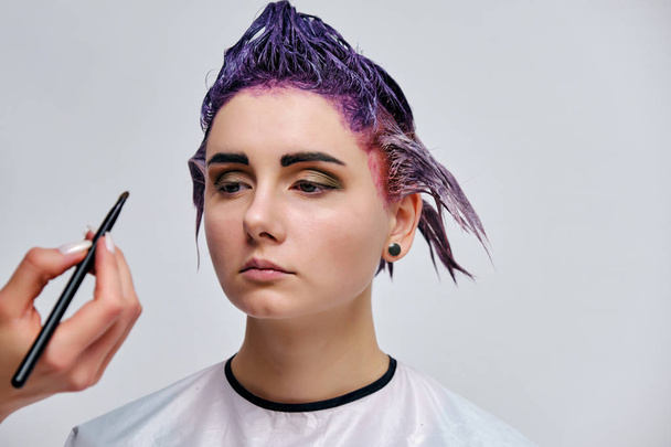 Beautiful girl with violet hair on white background master in make-up. Visible hand of the master and his makeup. The girl is in the middle of the frame and sits on a chair. - Foto, immagini