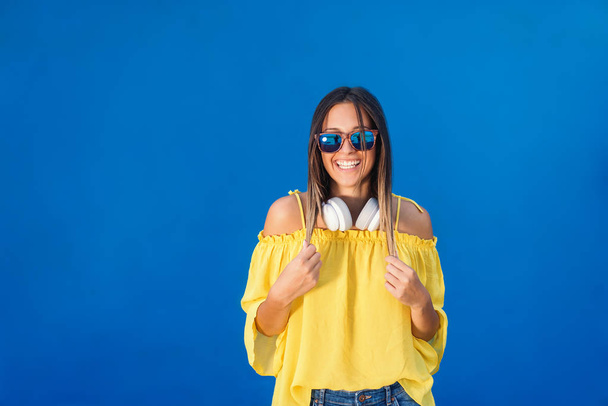 schöne Brünette in gelber Bluse, mit Sonnenbrille und Kopfhörer um den Hals posiert vor blauem Hintergrund. - Foto, Bild