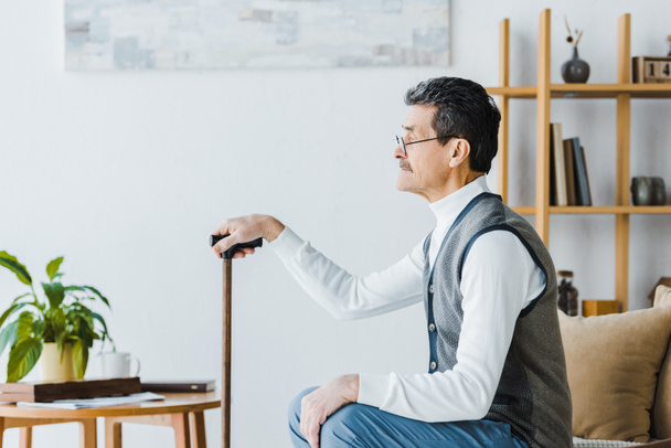  Trauriger Senior mit Schnurrbart sitzt und hält Spazierstock zu Hause - Foto, Bild