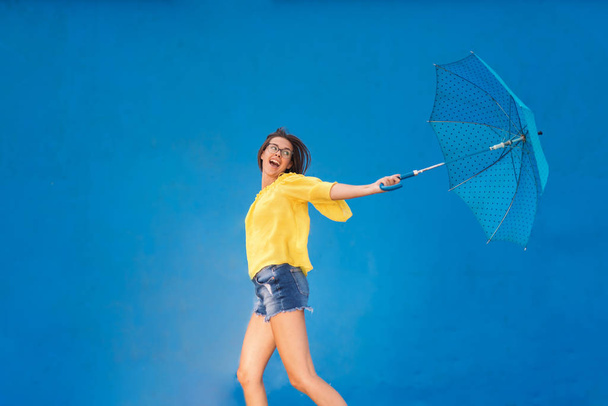 Aranyos tizenéves lány, barna haj, és szemüveget viselt sárga blúz és farmer rövidnadrág-kék esernyő, fekete pöttyökkel futás közben. Kék háttér. - Fotó, kép
