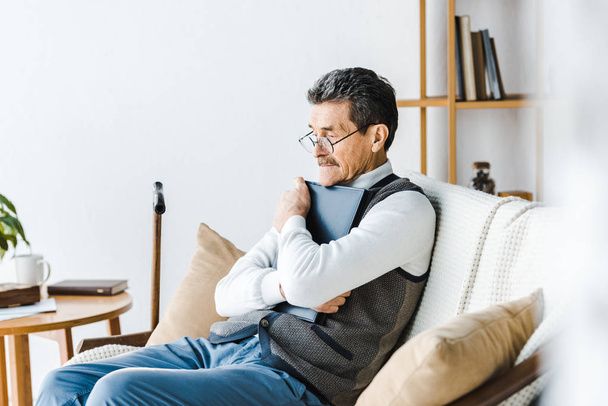 senior man in glasses hugging photo album while sitting on sofa - Foto, Imagen