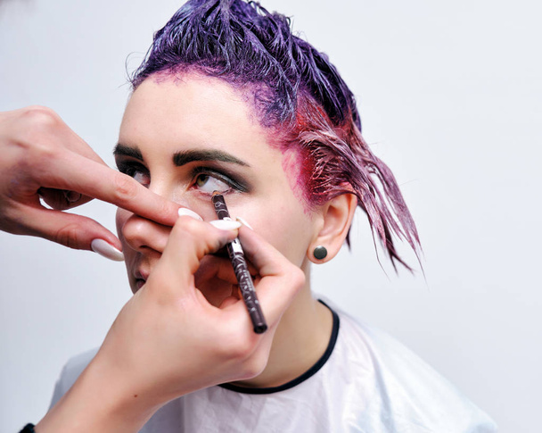 Beautiful girl with violet hair on white background master in make-up. Visible hand of the master and his makeup. The girl is in the middle of the frame and sits on a chair. - Фото, зображення