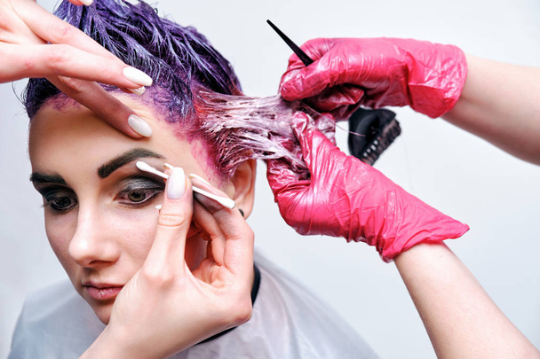 Beautiful girl with violet hair on white background master in make-up. Visible hand of the master and his makeup. The girl is in the middle of the frame and sits on a chair. - Fotoğraf, Görsel