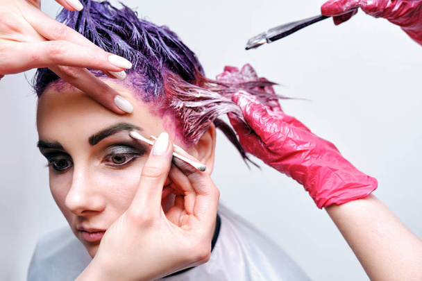 Beautiful girl with violet hair on white background master in make-up. Visible hand of the master and his makeup. The girl is in the middle of the frame and sits on a chair. - Zdjęcie, obraz