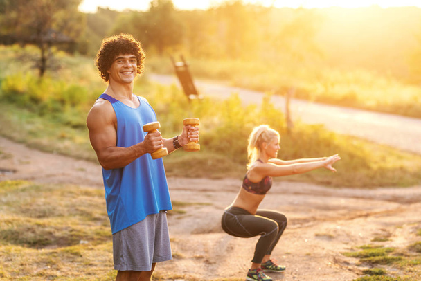 Hymyilevä lihaksikas mies urheiluvaatteissa tekee harjoituksia käsipainoilla luonnossa aurinkoisena kesäpäivänä. Taustalla nainen lämpenee
. - Valokuva, kuva
