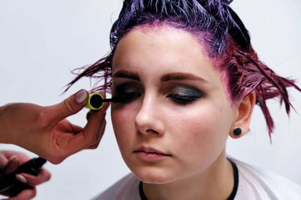 Beautiful girl with violet hair on white background master in make-up. Visible hand of the master and his makeup. The girl is in the middle of the frame and sits on a chair. - 写真・画像