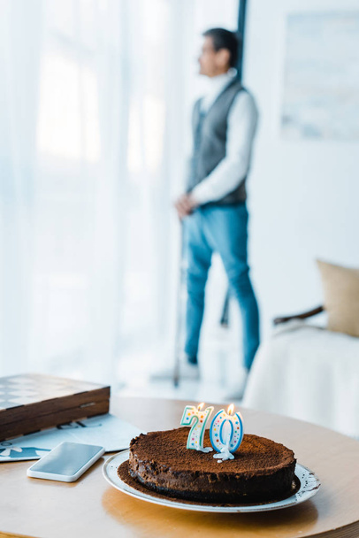 enfoque selectivo de pastel de cumpleaños con velas encendidas con hombre mayor solitario de pie con bastón de caminar en el fondo
 - Foto, Imagen