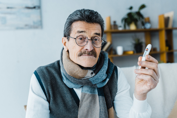 Kranker Senior im Schal hält Thermometer zu Hause - Foto, Bild