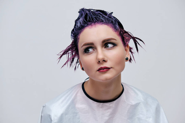 Beautiful girl with violet hair on white background master in make-up. Visible hand of the master and his makeup. The girl is in the middle of the frame and sits on a chair. - Fotografie, Obrázek