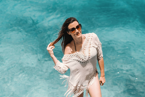 Hermosa mujer caucásica con cabello castaño largo, gafas de sol y en vestido de verano posando en el muelle. En el agua de fondo
. - Foto, imagen