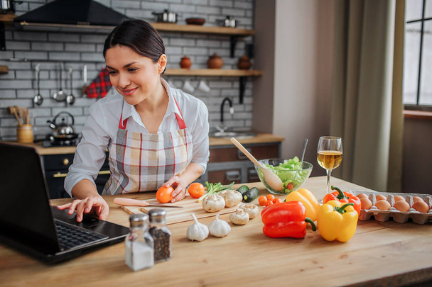 Cheerful adult woman sit at table in kitchen. She typing on keyboard and look on screen. Woman cook food. - Photo, image