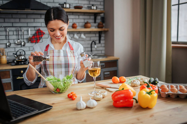 Cheerful nice woman sit at table in kitchen and put spices in bowl with salad. She looks happy. Woman wears apron. Colorful vegetables and eggs stand on table. - Foto, immagini