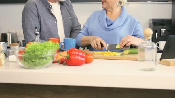 Pensioner couple preparing side dish together. Wife cutting cucumber and giving peace to the husband. - Séquence, vidéo