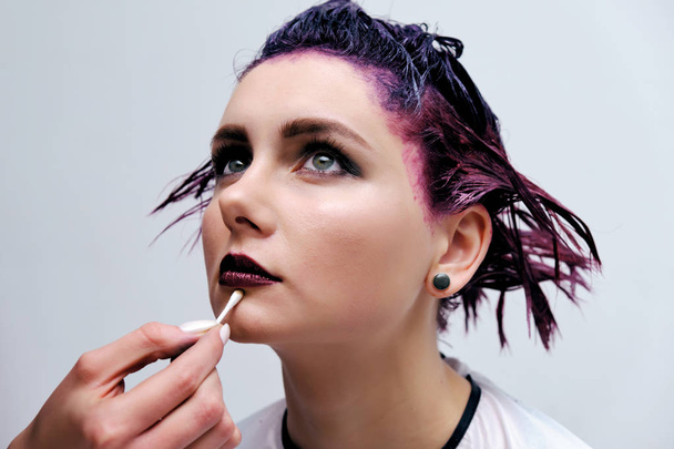 Beautiful girl with violet hair on white background master in make-up. Visible hand of the master and his makeup. The girl is in the middle of the frame and sits on a chair. - Fotografie, Obrázek