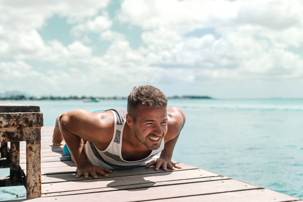 Handsome Caucasian unshaven man doing push ups on dock. In background ocean. - Foto, Imagen